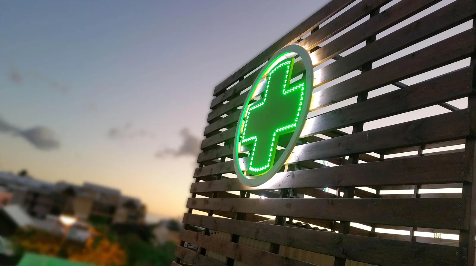 Une croix de pharmacie à LED vertes sur un bardage en bois, au coucher du soleil